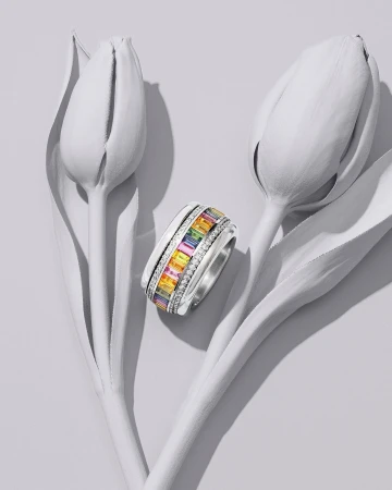 Close-up of a silver ring with multicolored gemstones next to two closed, white tulip blossoms on a light gray surface.