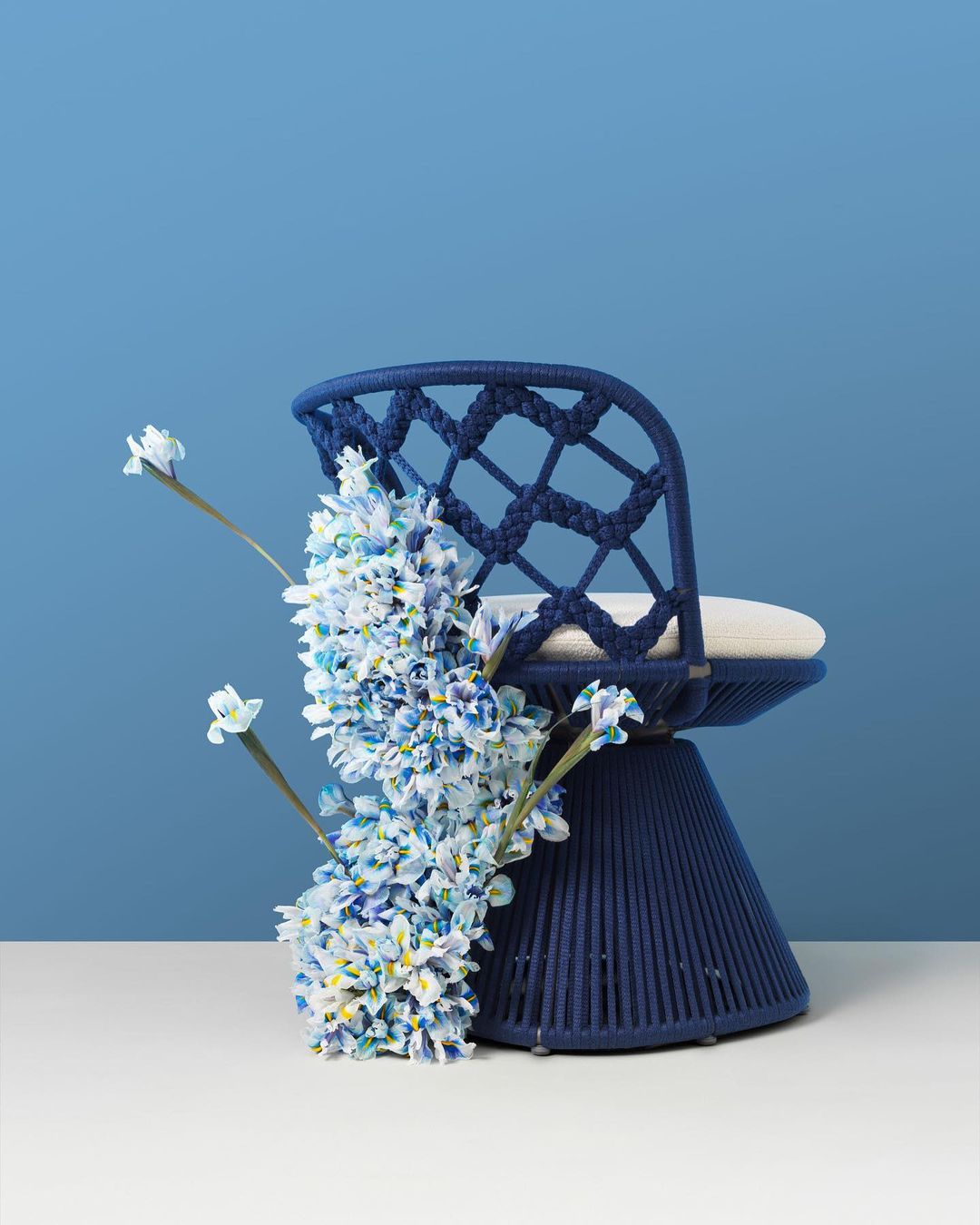 Navy blue woven chair with off-white cushion, adorned with white and blue flowers, against a gradient blue backdrop for interior design.