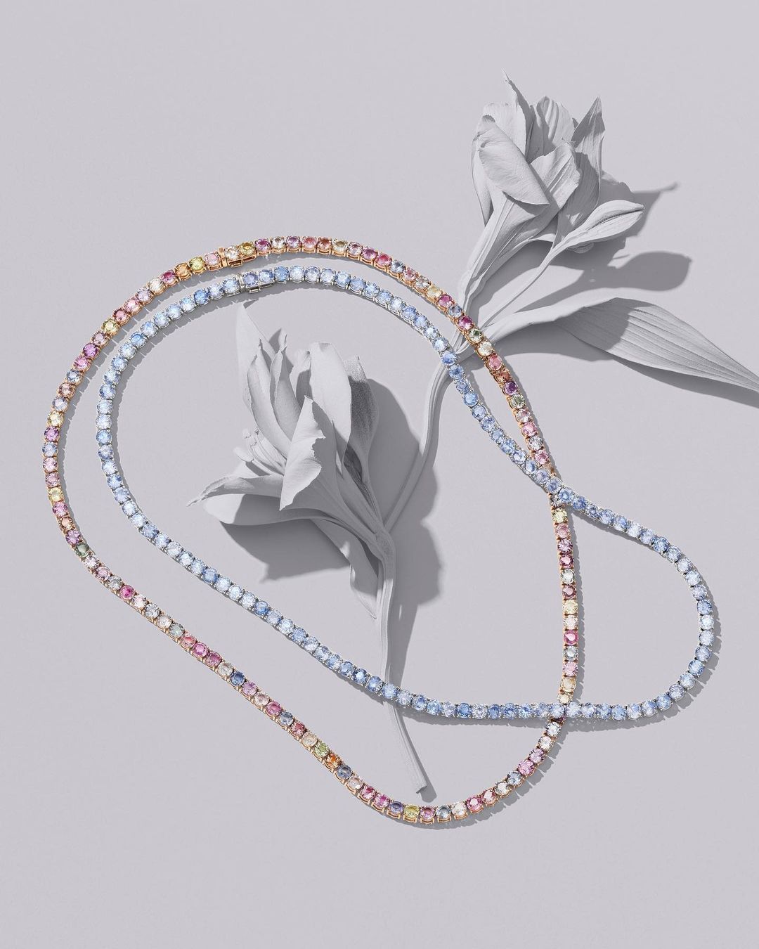 Elegant jewelry flat lay showcases two multi-color gemstone necklaces draped beside elegant white flowers on a soft grey background.
