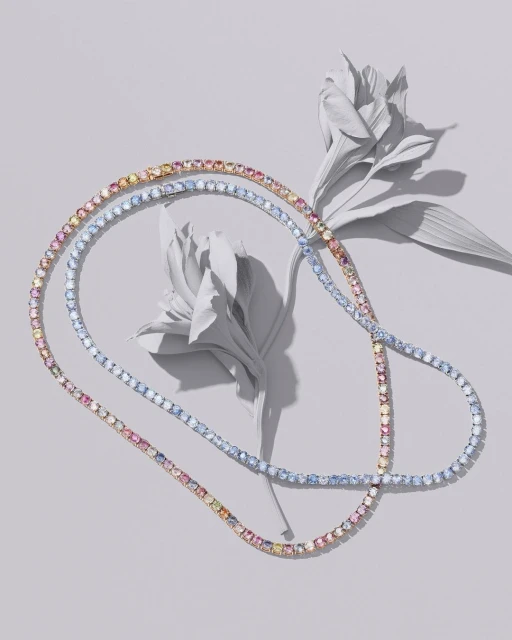 Elegant jewelry flat lay showcases two multi-color gemstone necklaces draped beside elegant white flowers on a soft grey background.