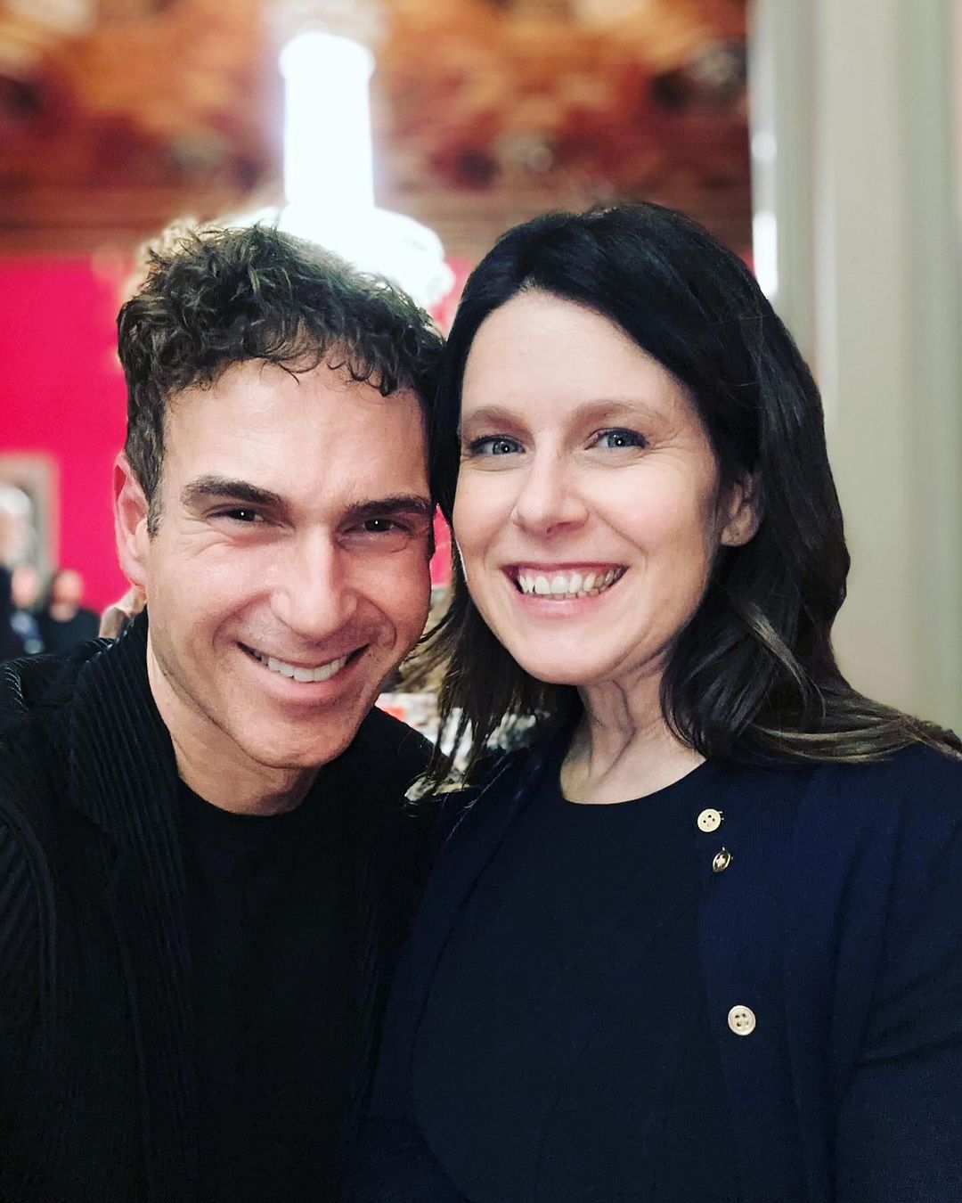 Close-up portrait of a smiling man and woman posing indoors, with a decorated ceiling and wall visible in the background.
