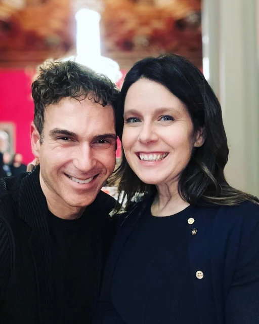 Close-up portrait of a smiling man and woman posing indoors, with a decorated ceiling and wall visible in the background.