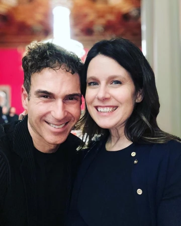 Close-up portrait of a smiling man and woman posing indoors, with a decorated ceiling and wall visible in the background.