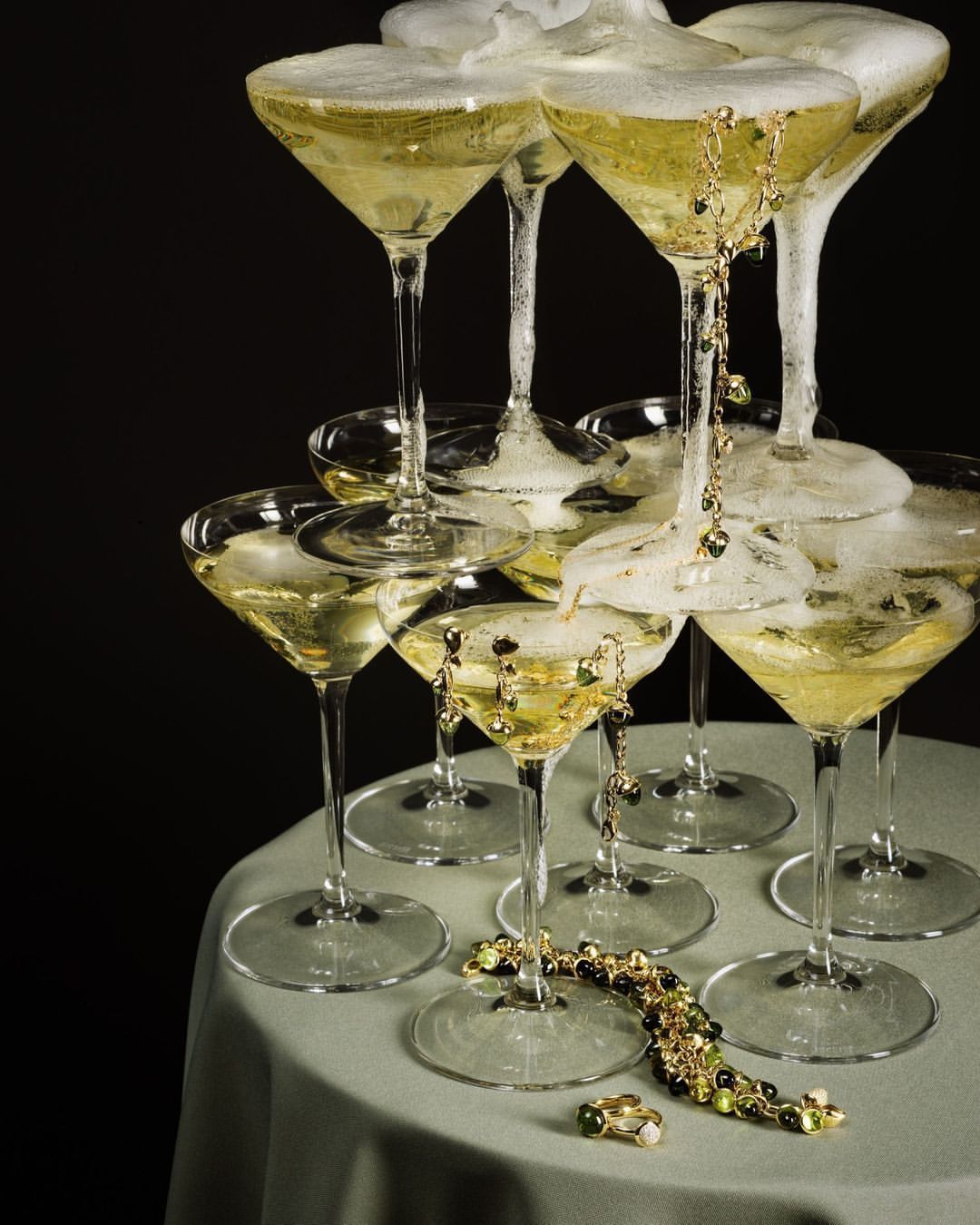 Champagne tower of martini glasses with golden jewelry draped, set on a round table against a dark backdrop, celebratory vibe.