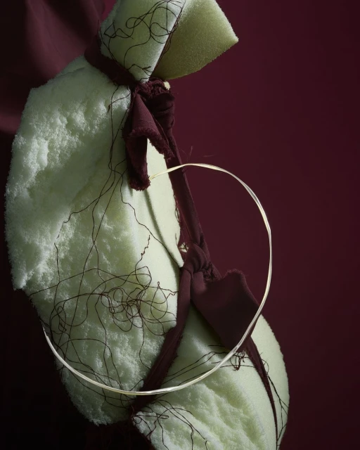 Abstract still life: light-green textile draped over a maroon fabric, a thin metallic hoop rests between the layers of textile.