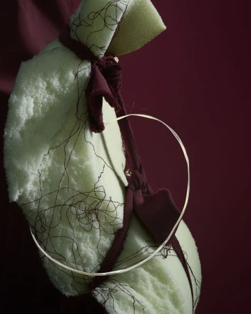 Abstract still life: light-green textile draped over a maroon fabric, a thin metallic hoop rests between the layers of textile.