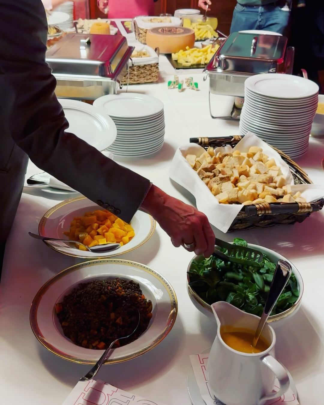 Food service table laden with various salads, breads, cheeses, and hot dishes, set up for a self-serve buffet-style event.