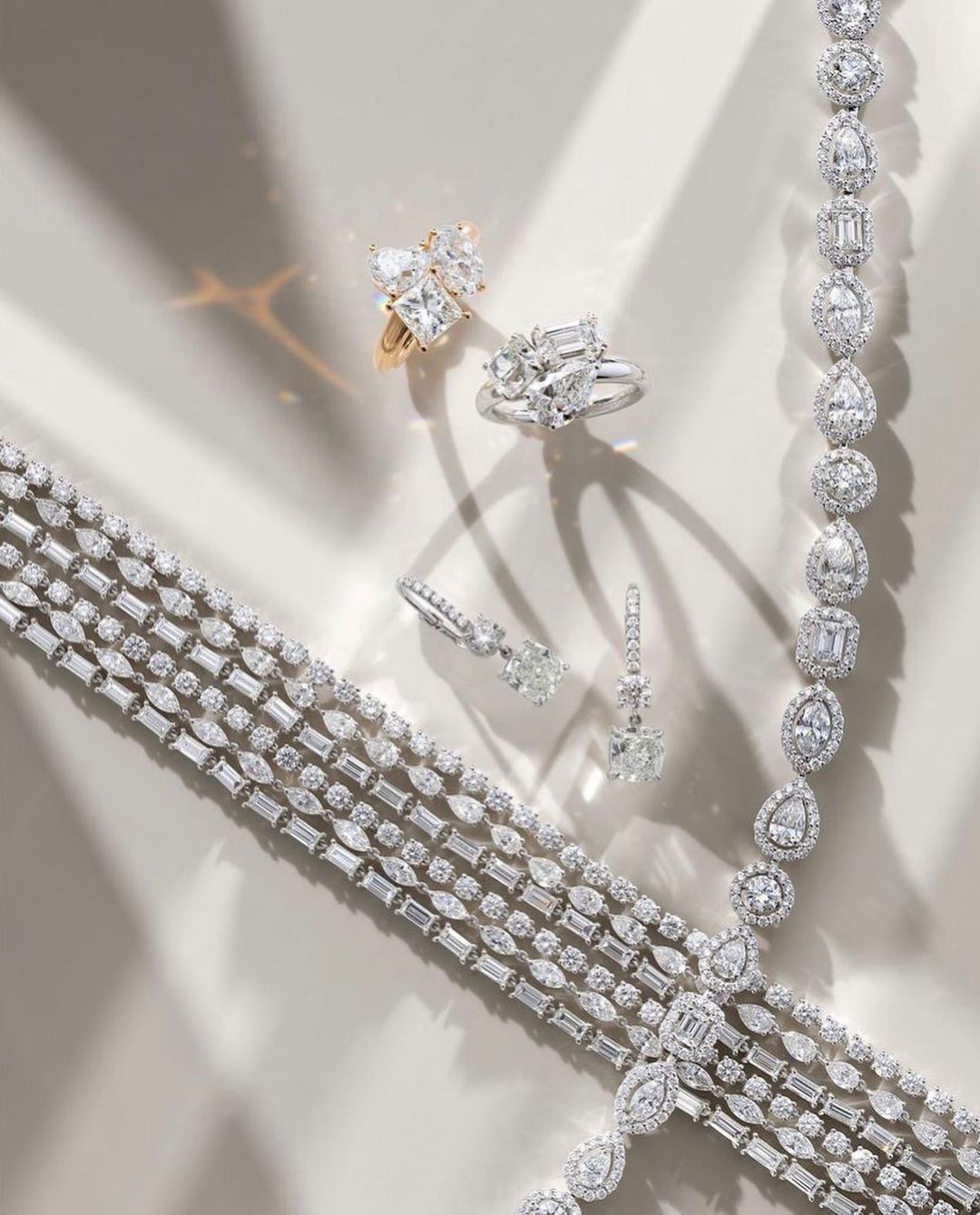 High-angle shot of diamond rings, earrings, bracelets, and necklaces displayed on a neutral surface, with light creating soft shadows.