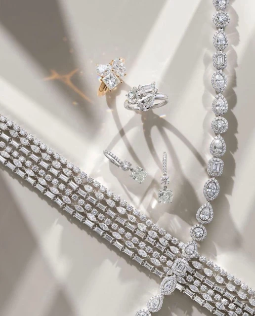 High-angle shot of diamond rings, earrings, bracelets, and necklaces displayed on a neutral surface, with light creating soft shadows.
