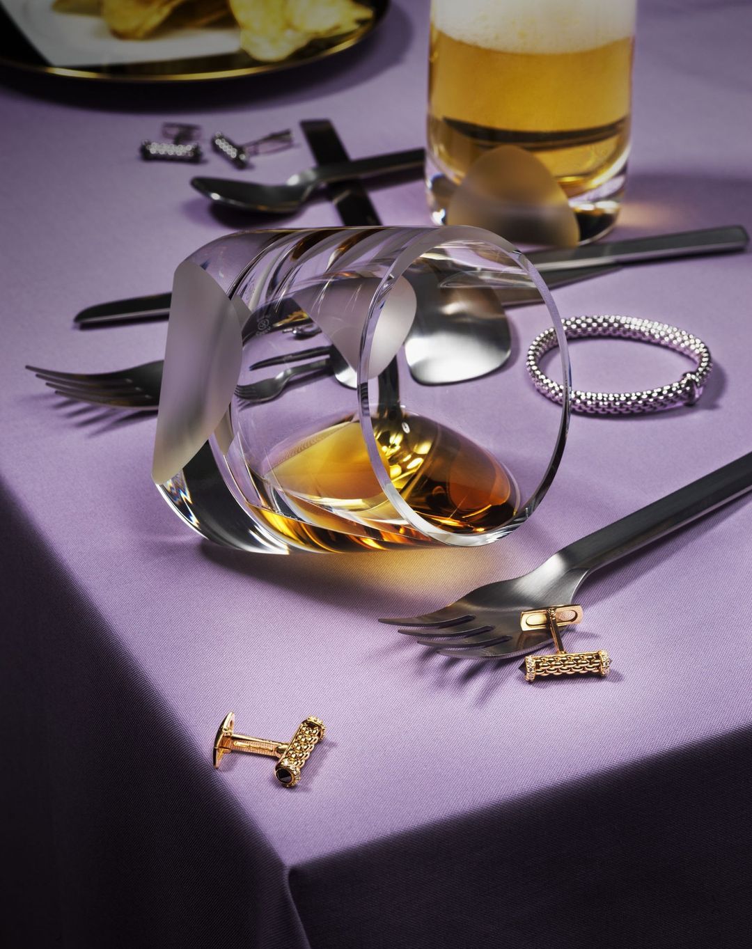 Crystal glass with amber liquor on a lilac linen surface, next to golden cufflinks, beer, silverware, and potato chips.