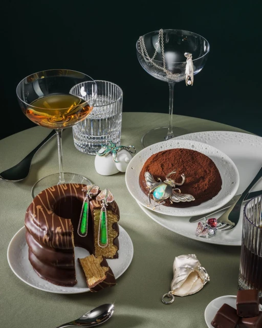 Still life featuring gourmet desserts, jewelry, and crystal glasses on a pale green table against a dark green background.