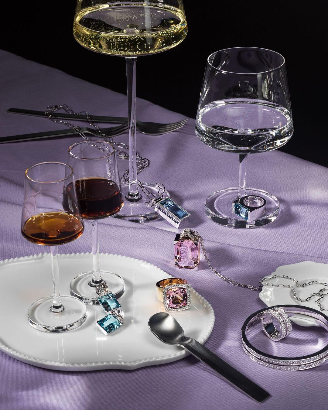 Close-up of highball glasses with clear and amber liquors alongside assorted silver jewelry on a white tray and lavender tablecloth.
