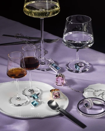 Close-up of highball glasses with clear and amber liquors alongside assorted silver jewelry on a white tray and lavender tablecloth.