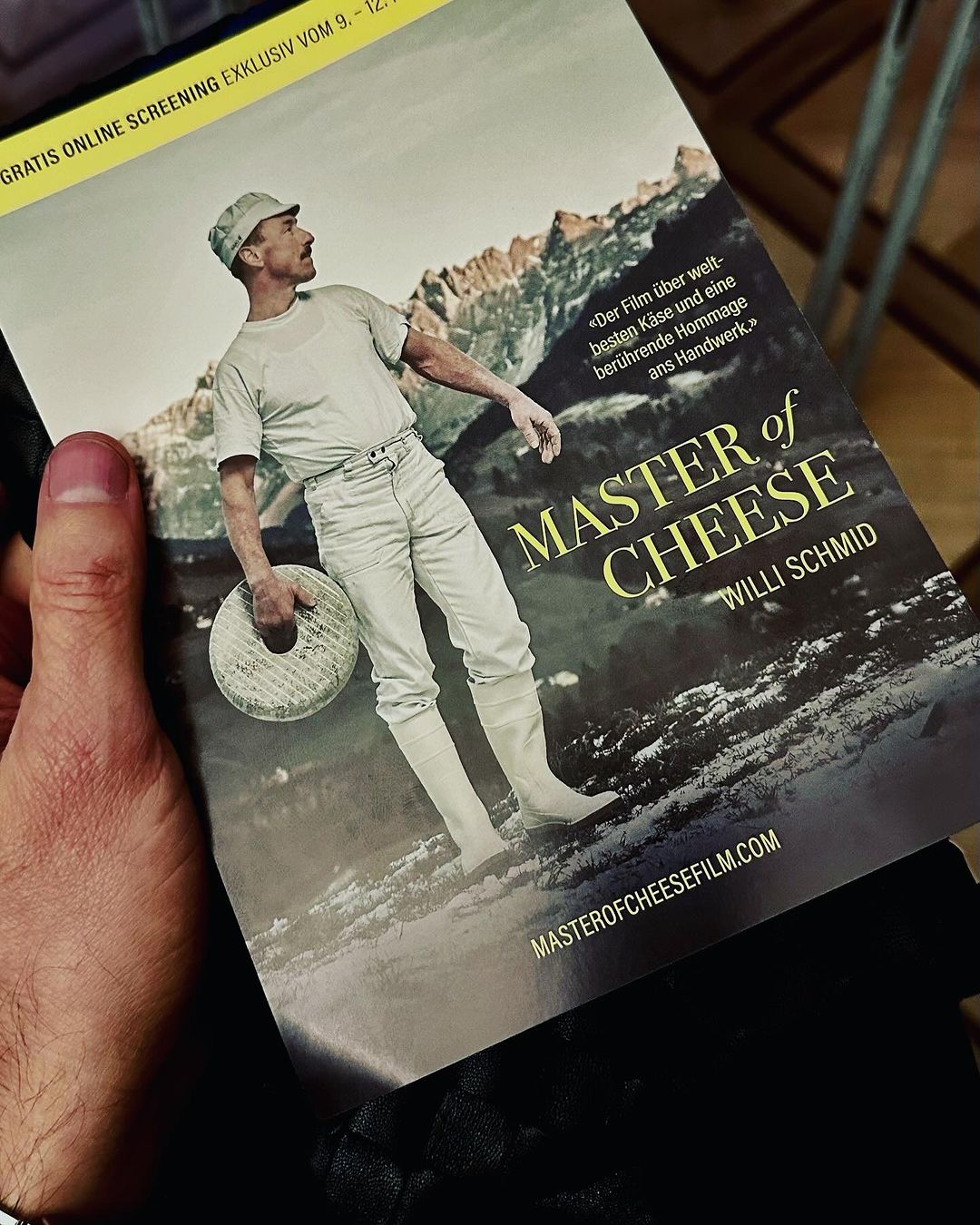 A person holding a promotional flyer for the film Master of Cheese featuring a man in historical attire holding a cheese wheel against a mountain backdrop.