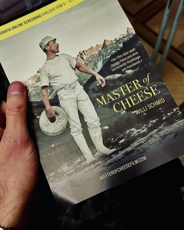 A person holding a promotional flyer for the film Master of Cheese featuring a man in historical attire holding a cheese wheel against a mountain backdrop.