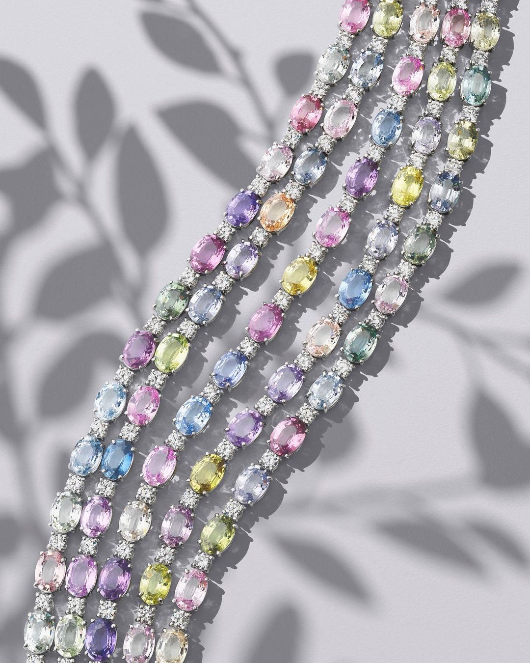 Overhead studio shot displays five delicate gemstone bracelets with colorful pastel gems and diamond accents on a pale gray surface.