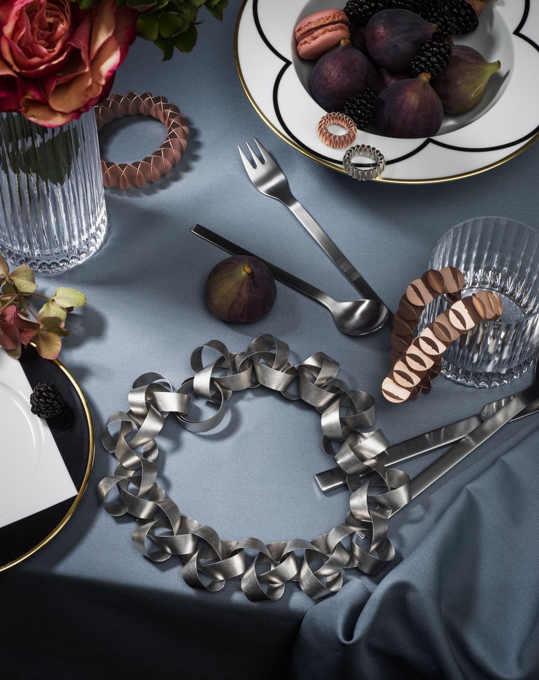 Still life of figs, blackberries, macarons on a plate with silverware and decorative objects arranged on a gray-blue cloth surface.