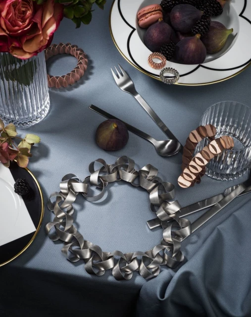 Still life of figs, blackberries, macarons on a plate with silverware and decorative objects arranged on a gray-blue cloth surface.