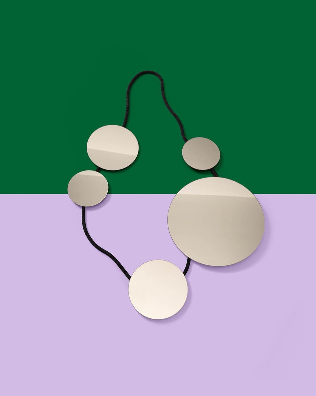 Abstract product shot of a modern necklace with round, flat pendants connected by a black cord against a green and lavender background.