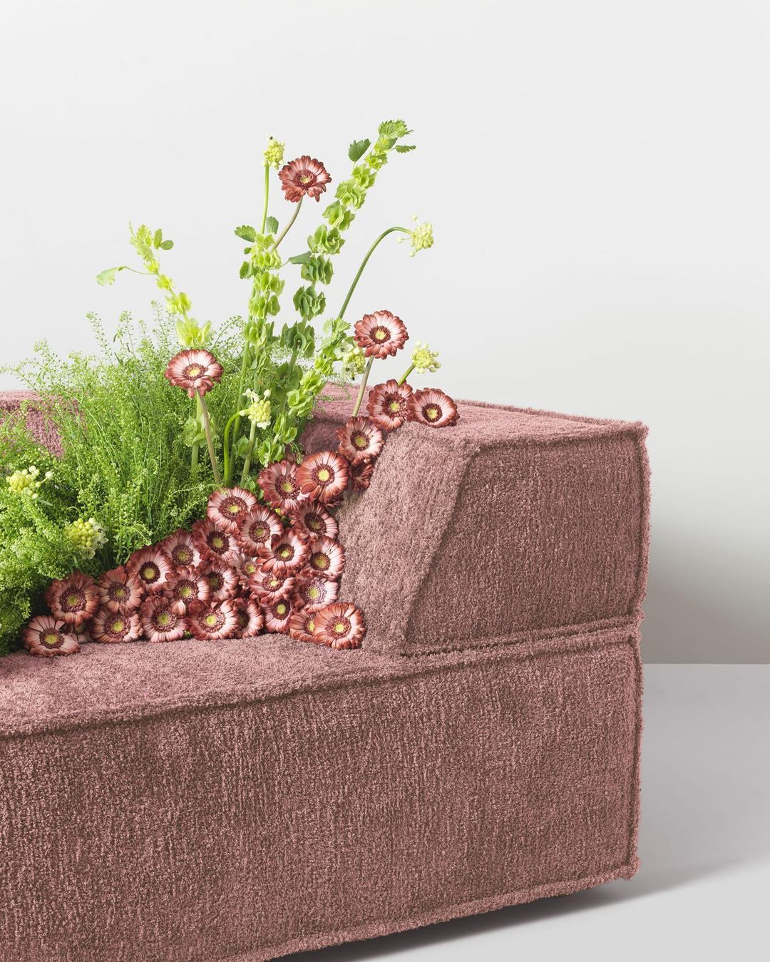 Modern mauve couch topped with wildflowers in studio setting. Close-up view, soft lighting, and neutral white background.
