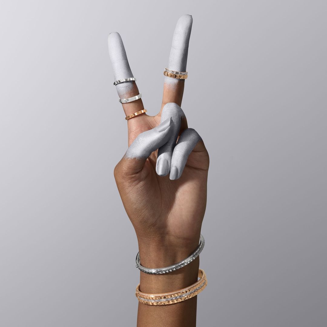 Hand painted silver making a peace sign with jewelry including rings and bracelets against a grey backdrop in a studio.