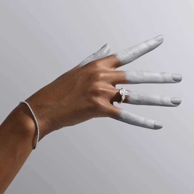 Elegant diamond ring on a hand with silver paint, alongside a delicate diamond bracelet, jewelry product shot on neutral grey.