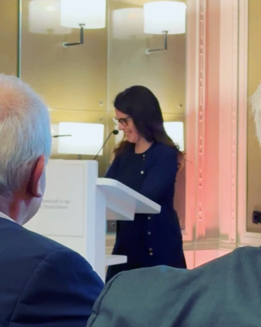 Adult woman with dark curly hair wearing glasses and a dark blue dress stands at a white podium and speaks to an audience.