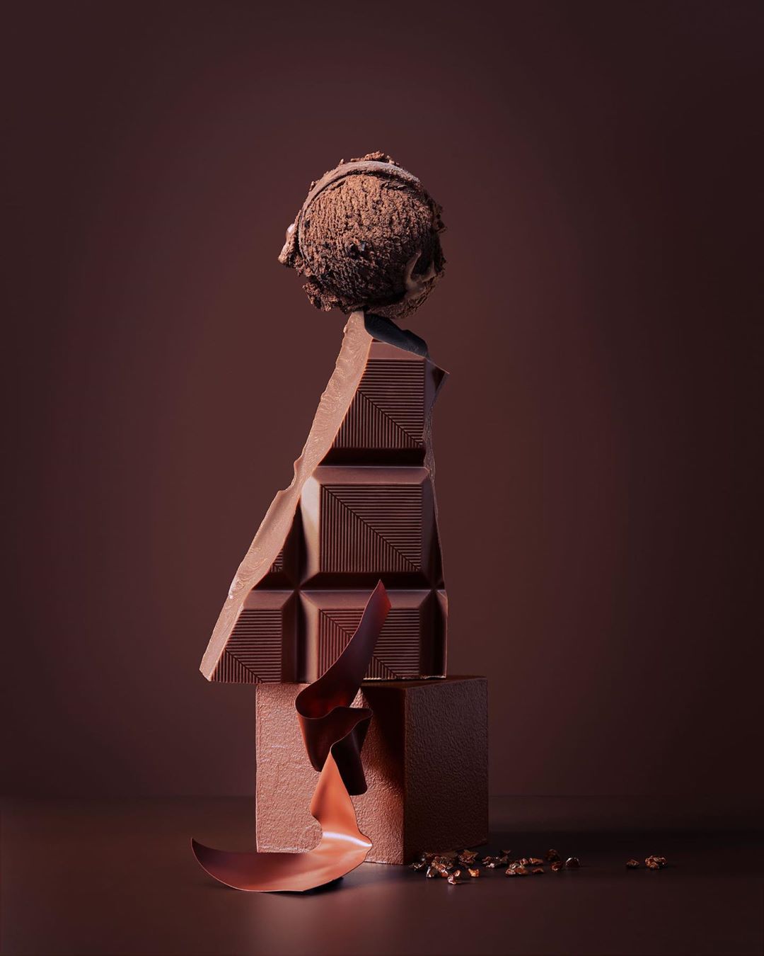 Still life of chocolate ice cream scoop balanced on a chunk of dark chocolate alongside chocolate shavings on a brown background.