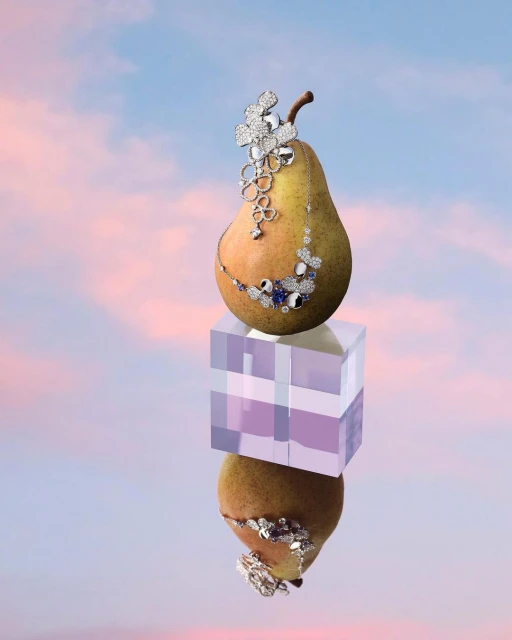 Pear adorned with jewelry, perched on a translucent cube with mirrored reflection against a soft pink and blue sky backdrop.