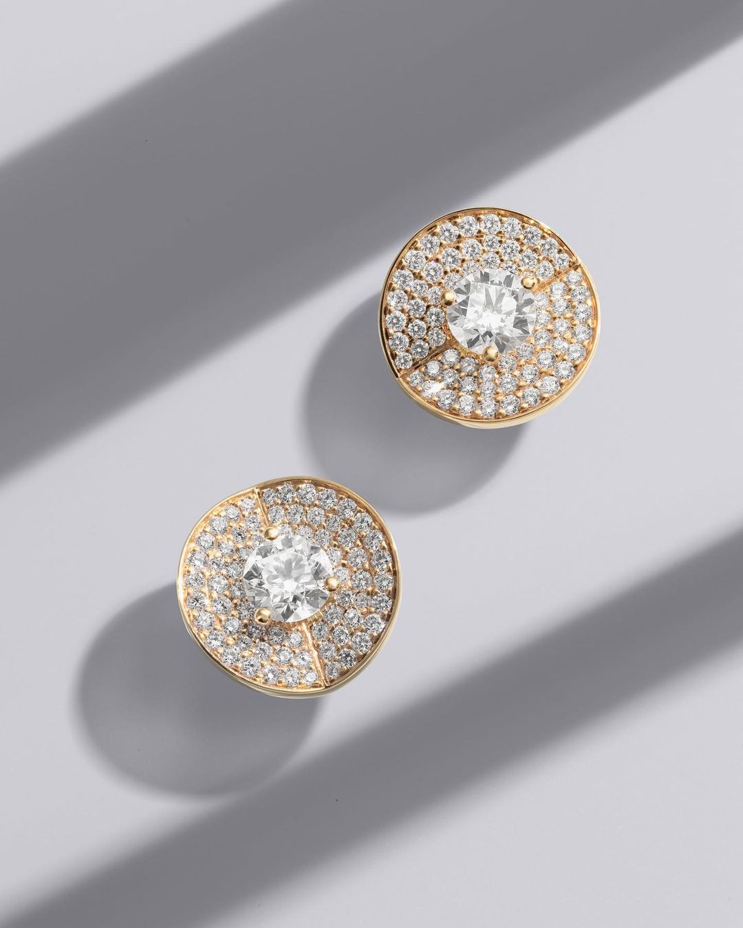 Close-up of two gold stud earrings with pave diamonds surrounding a central larger diamond, on a light grey background with shadows.