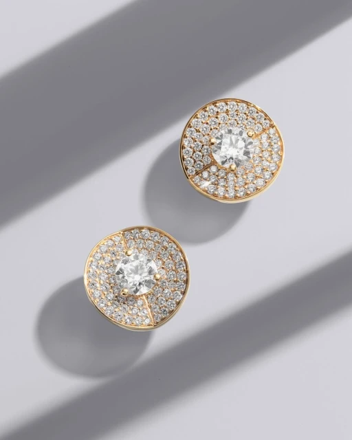 Close-up of two gold stud earrings with pave diamonds surrounding a central larger diamond, on a light grey background with shadows.