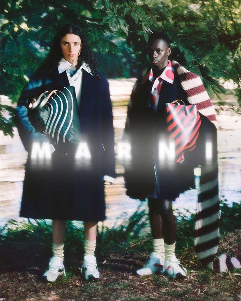 Fashion shot of two models standing in a wooded outdoor setting, showcasing patterned bags and coats by the brand Marni. 