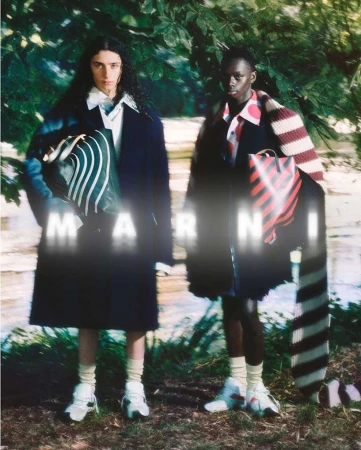 Fashion shot of two models standing in a wooded outdoor setting, showcasing patterned bags and coats by the brand Marni. 