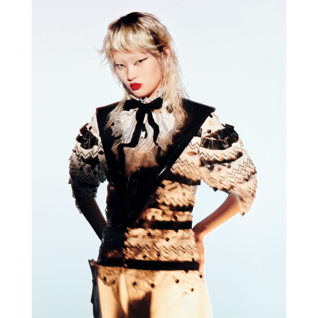 Blonde-haired model posing in a fashion editorial image with a decorative sheer top and dark vest against a pale blue background.