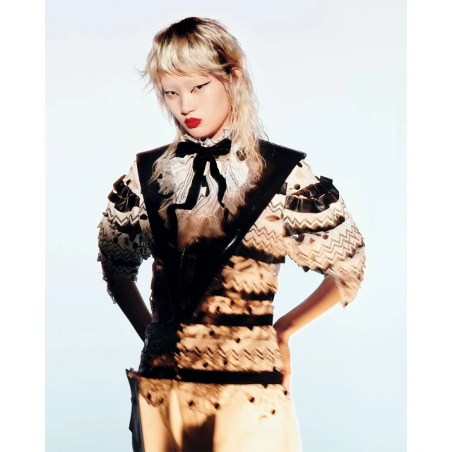 Blonde-haired model posing in a fashion editorial image with a decorative sheer top and dark vest against a pale blue background.