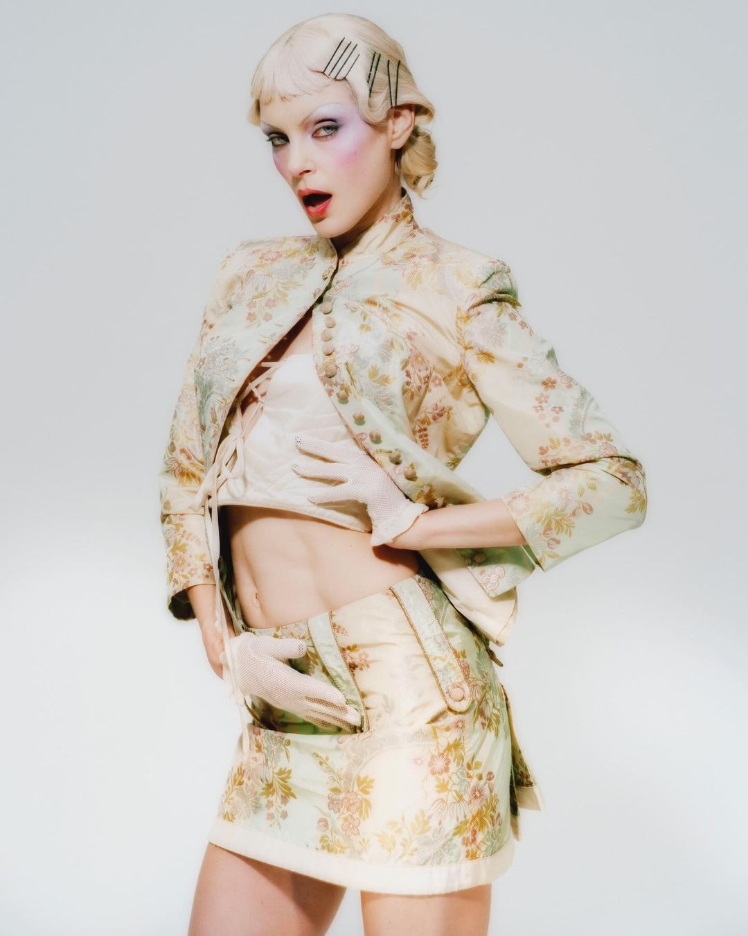 Young woman in pale floral brocade miniskirt suit and gloves poses in a photo studio against a light backdrop, wearing face makeup.