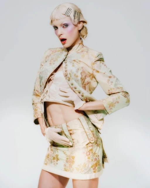 Young woman in pale floral brocade miniskirt suit and gloves poses in a photo studio against a light backdrop, wearing face makeup.