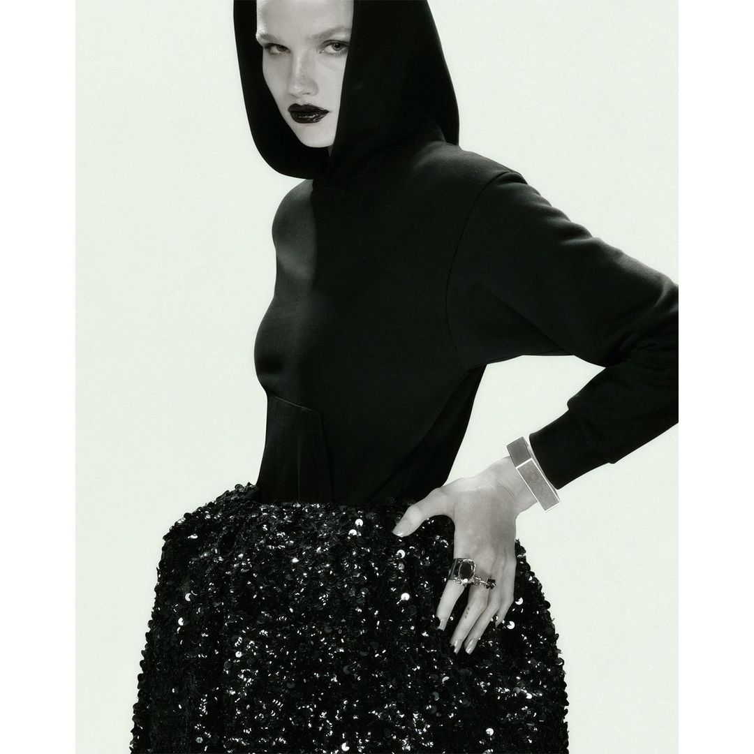 Monochrome fashion shot of a woman with dark lipstick wearing a hooded sweatshirt and a sequined skirt against a white backdrop.