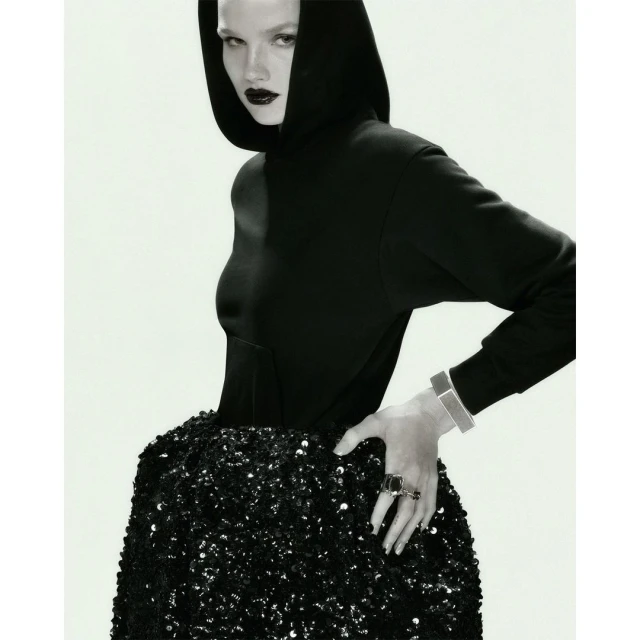 Monochrome fashion shot of a woman with dark lipstick wearing a hooded sweatshirt and a sequined skirt against a white backdrop.