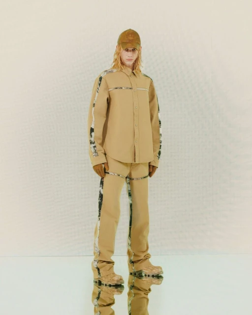 Tan outfit on a slender adult fashion model with long blond hair, wearing a baseball cap and boots, standing against a white background.