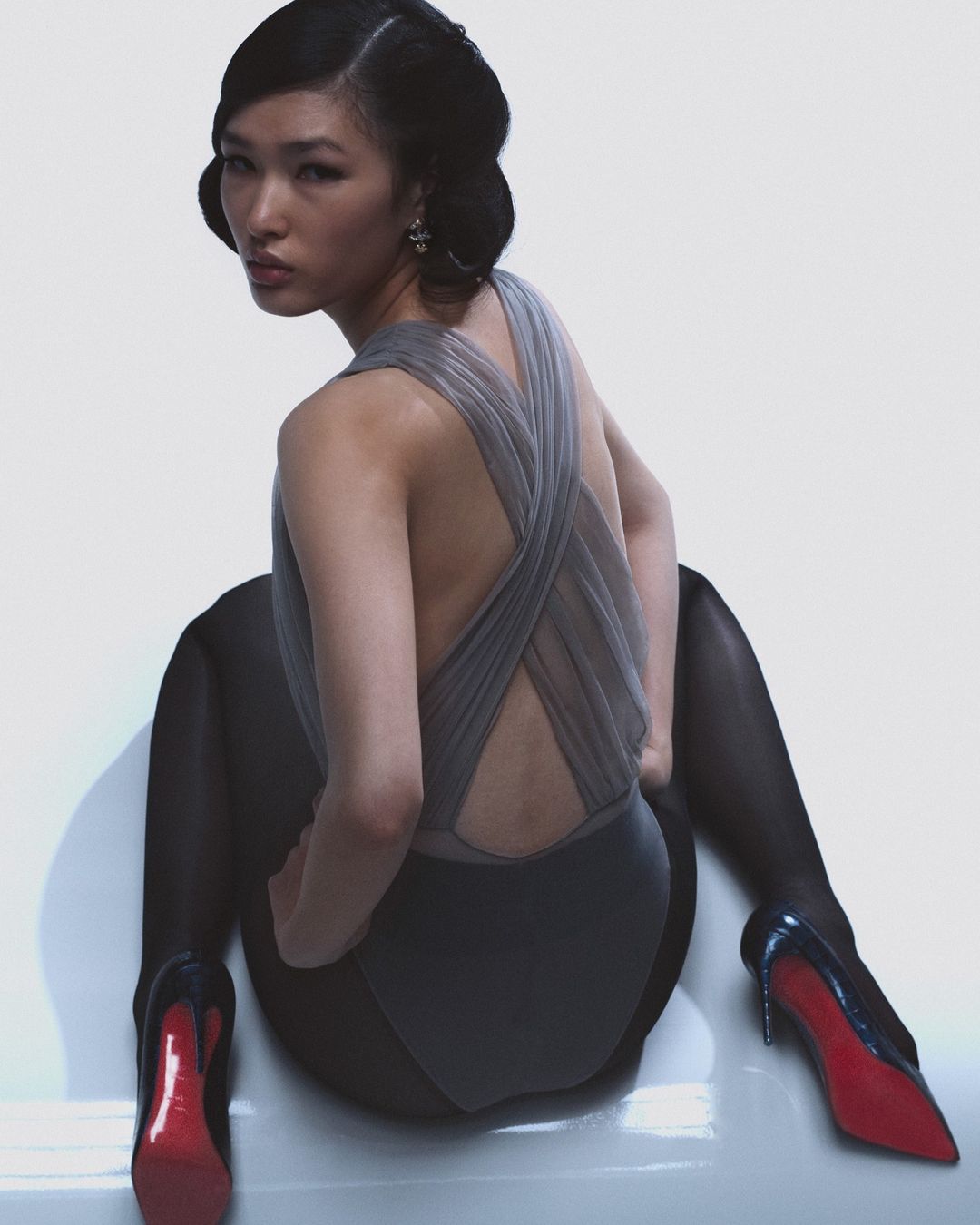 Elegant Asian woman with an updo, silver dress, black tights, and red high heels in a white photo studio setting. Fashion photography.