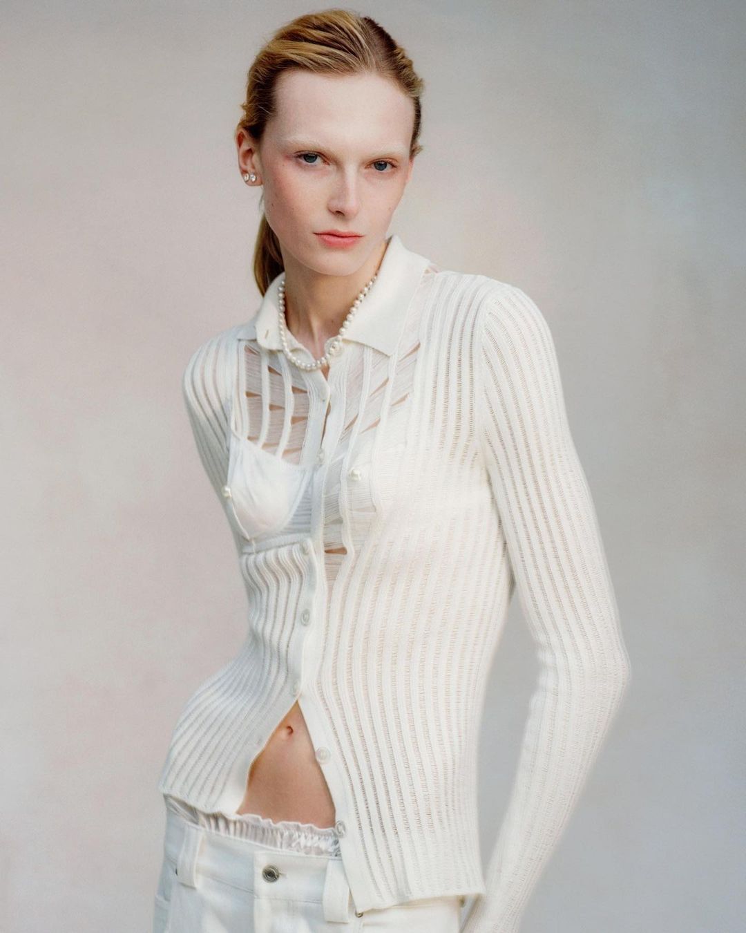 Blonde woman poses in a white cardigan and jeans against a neutral background in a studio shot. The image has a light and airy feel.