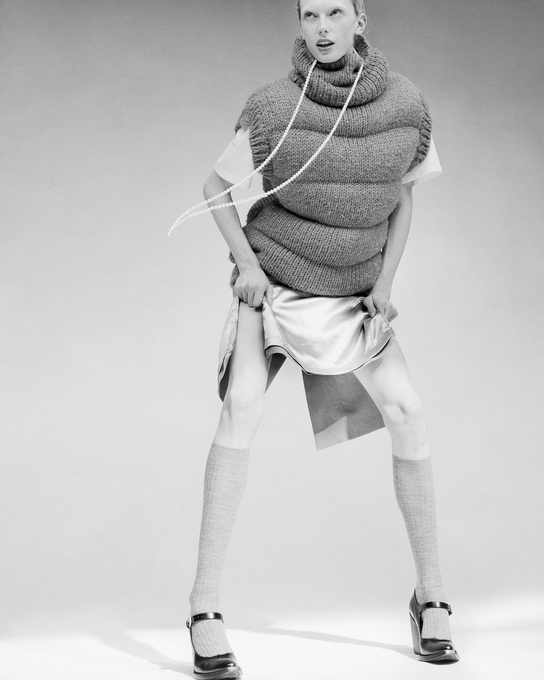 A woman stands in a photo studio, wearing a bulbous knit top, iridescent skirt, knee socks, and platform Mary Janes, with a pearl necklace.