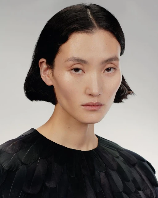 Headshot of a woman with dark, short hair wearing a black top covered in feathers against a neutral grey backdrop in a studio.