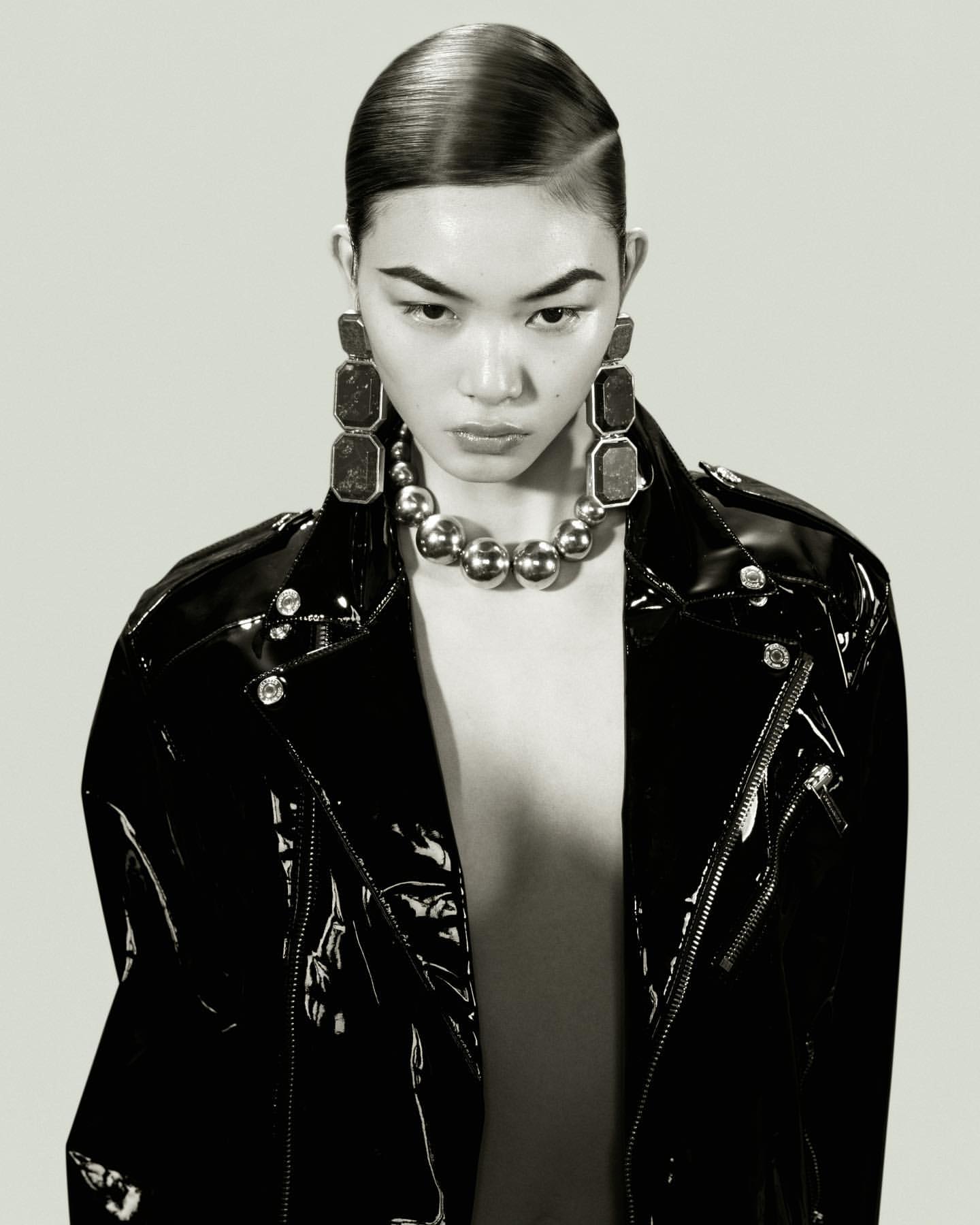Glamorous portrait of an Asian woman in a black patent leather jacket and statement jewellery, with sleek hair and a grey background.
