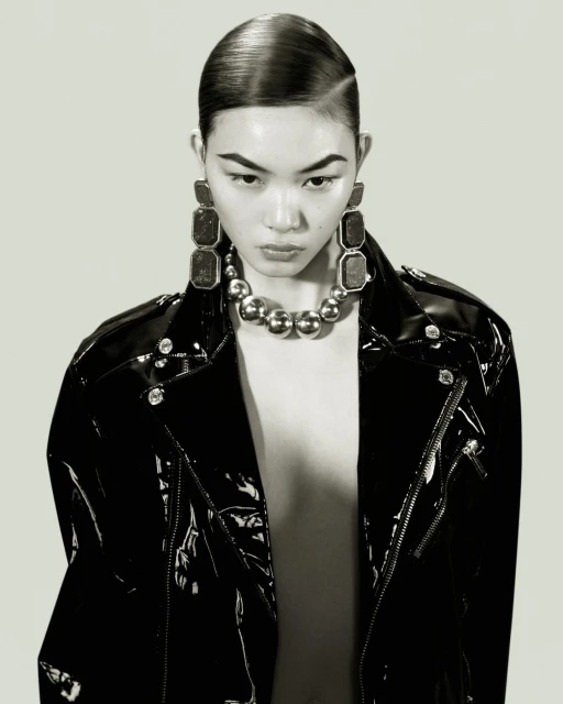 Glamorous portrait of an Asian woman in a black patent leather jacket and statement jewellery, with sleek hair and a grey background.