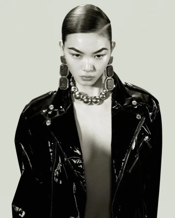 Glamorous portrait of an Asian woman in a black patent leather jacket and statement jewellery, with sleek hair and a grey background.