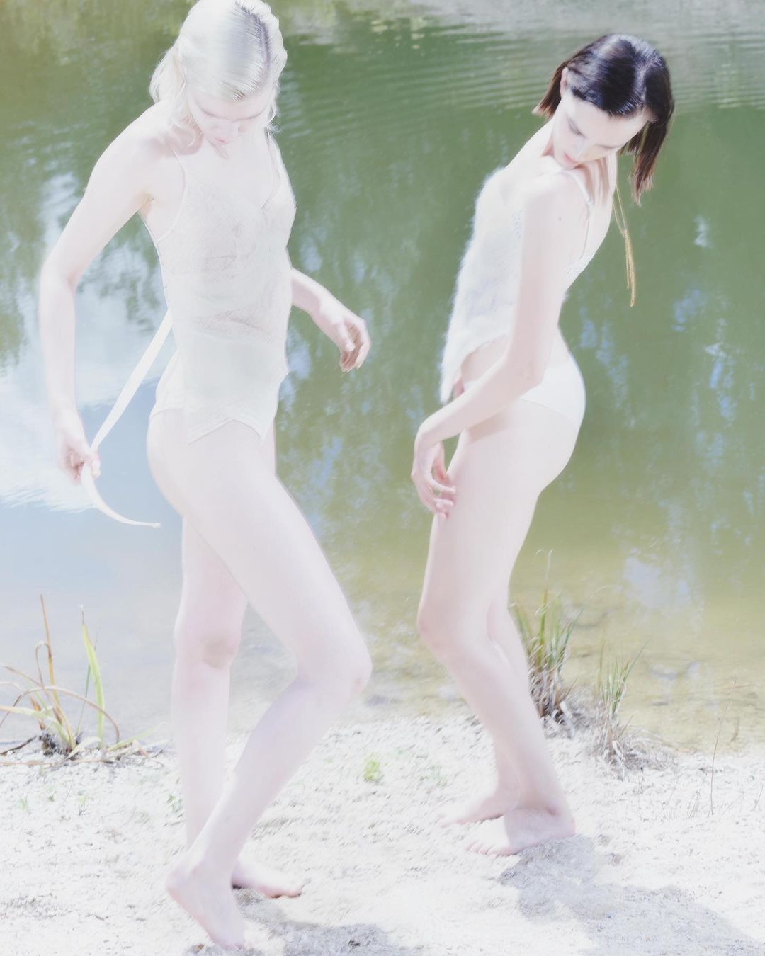 Two women in light-colored swimwear pose on a sandy bank near a green-tinged body of water on a bright, overcast day.