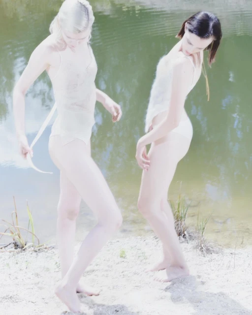 Two women in light-colored swimwear pose on a sandy bank near a green-tinged body of water on a bright, overcast day.