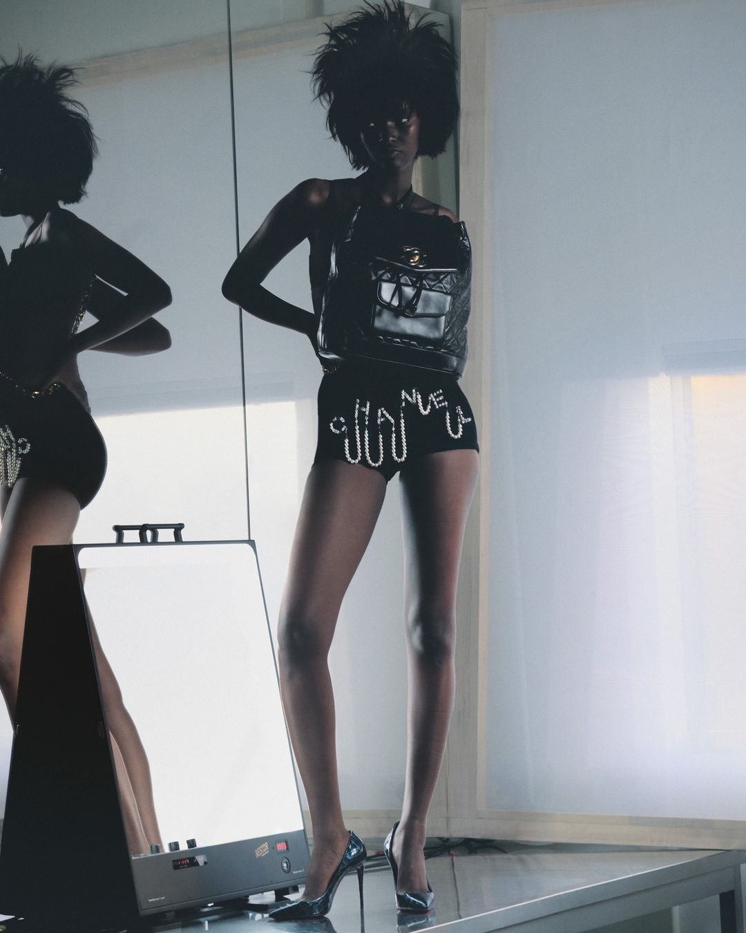 Model with voluminous dark hair wearing embellished black shorts and heels standing in a studio by Arnaud Lajeunie